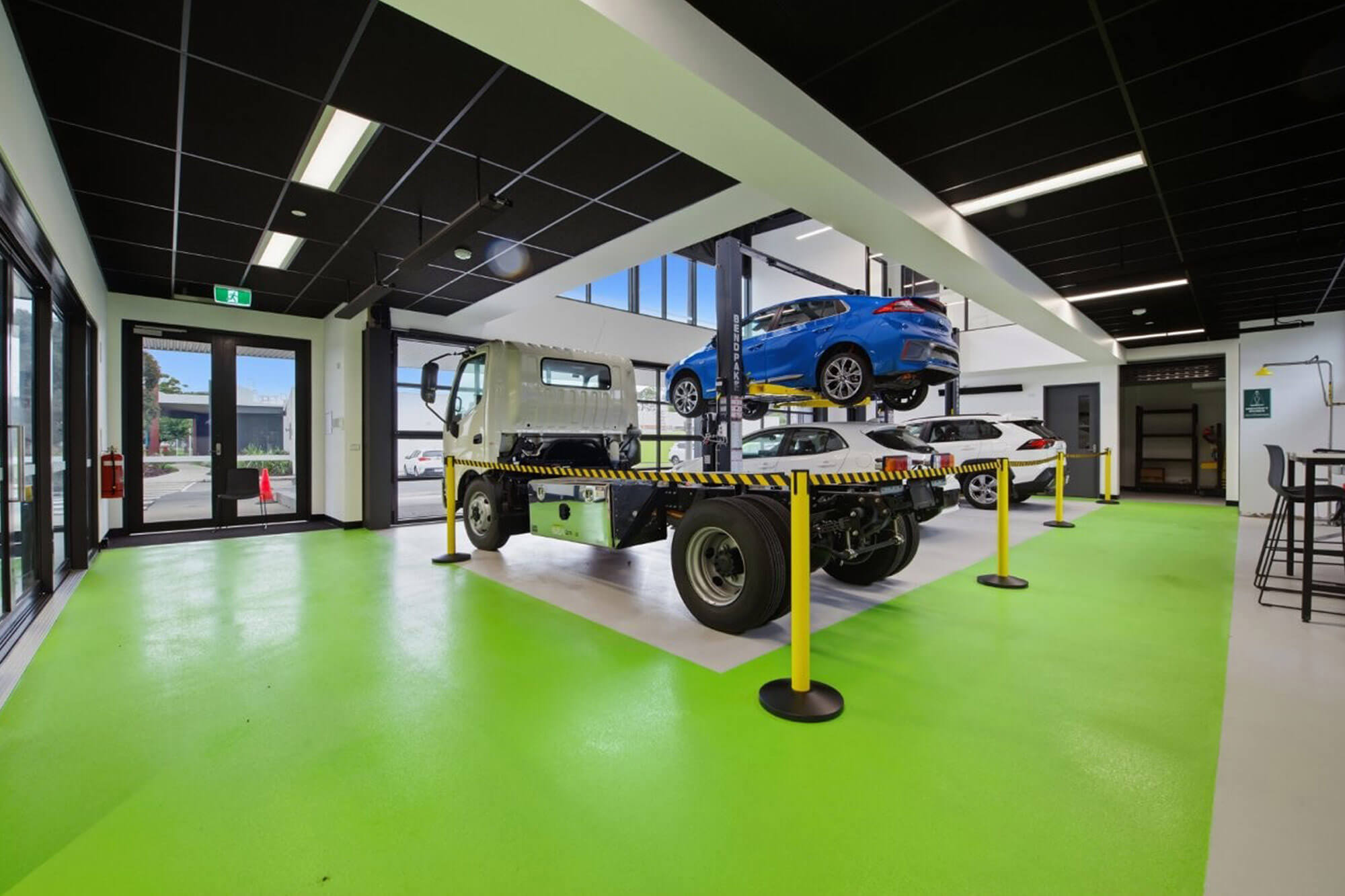 View inside the new TAFE Gippsland Clean Energy Centre showing lower floor with black ceiling, white walls and bright green concrete floor. Behind yellow and black safety line is a white truck cab, and two white cars, with a blue car raised on a hoist. They are electric vehicles.