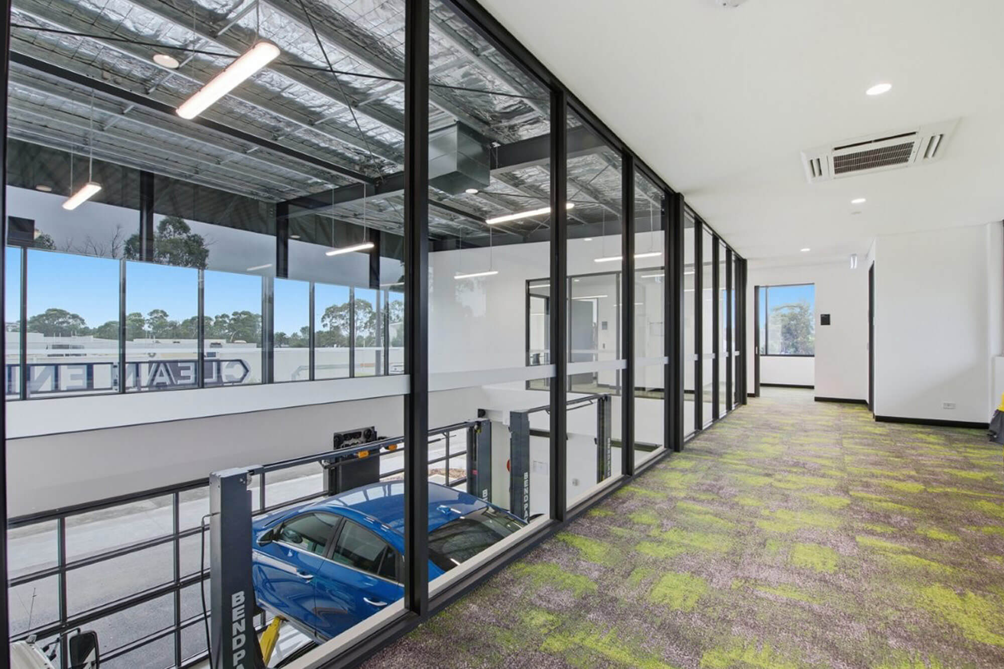 View from inside the new TAFE Gippsland Clean Energy Centre looking out windows down to the lower floor where there is an automotive area with a blue car raised on a hoist.